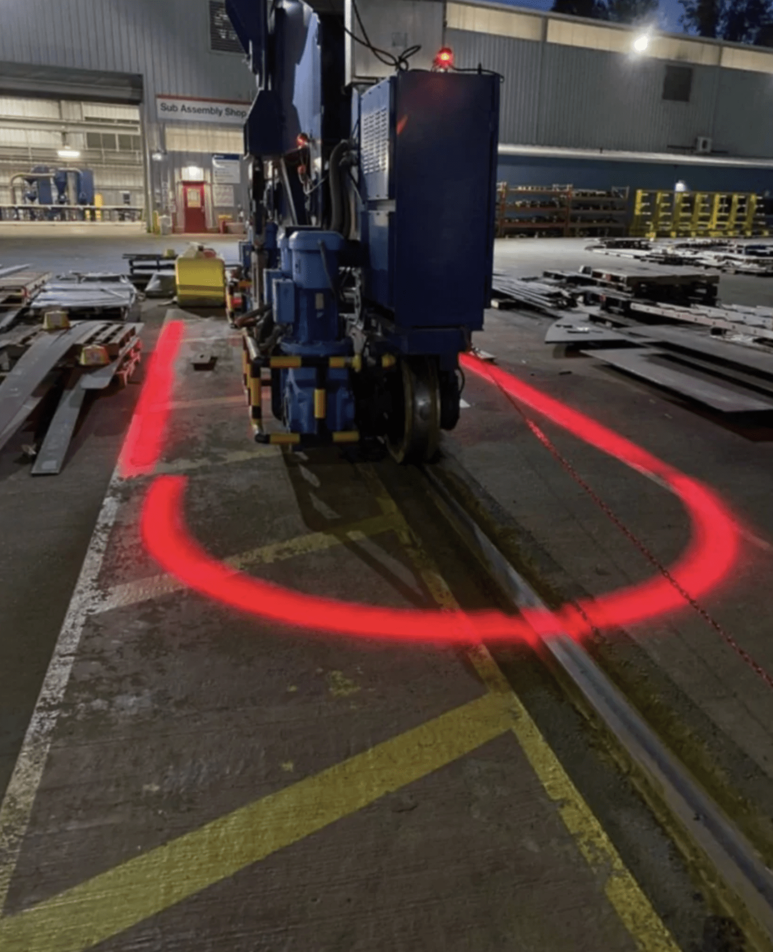 Industrial rail-mounted machine in a factory setting with red safety light projection outlining a curved exclusion zone around the vehicle.