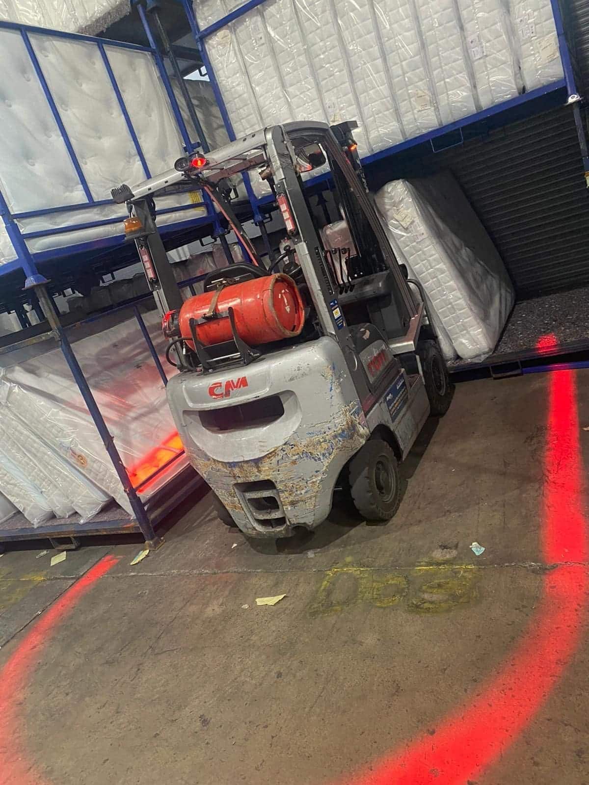 A forklift operating in a warehouse with plastic-wrapped mattresses, equipped with a red safety light system projecting a visible arc and lines on the floor to mark a safe distance zone.
