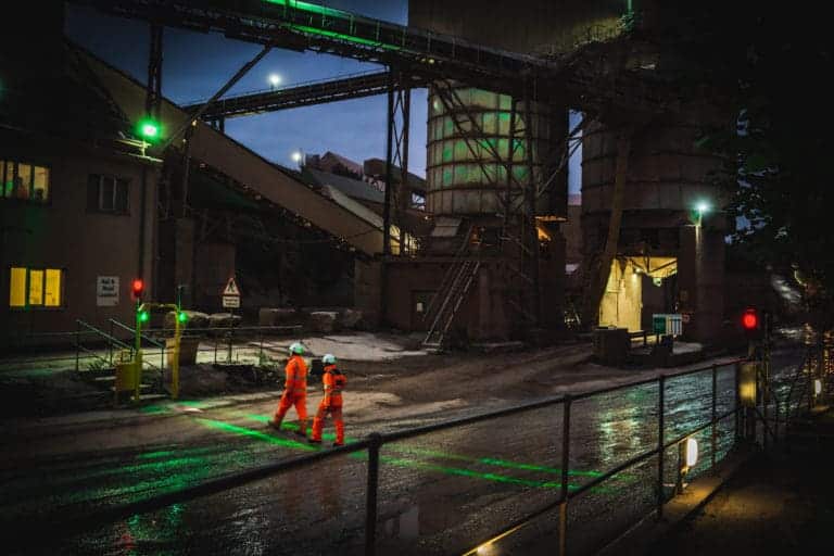 Two workers in high-visibility PPE cross a wet industrial site at night using a green-lit FHOSS safety crossing system, with overhead conveyors and illuminated buildings in the background.