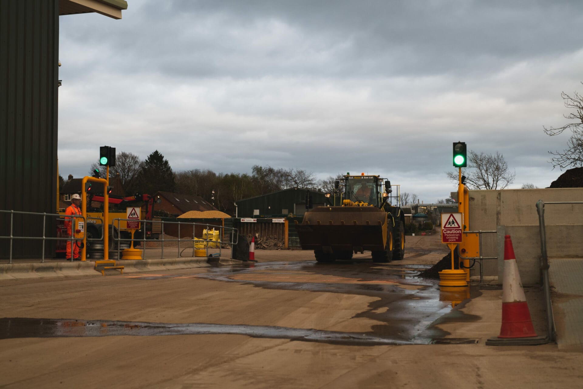 A construction site with a large front loader vehicle approaching a pedestrian crossing area, featuring green traffic lights, warning signs, and a worker in high-visibility clothing standing safely behind barriers.